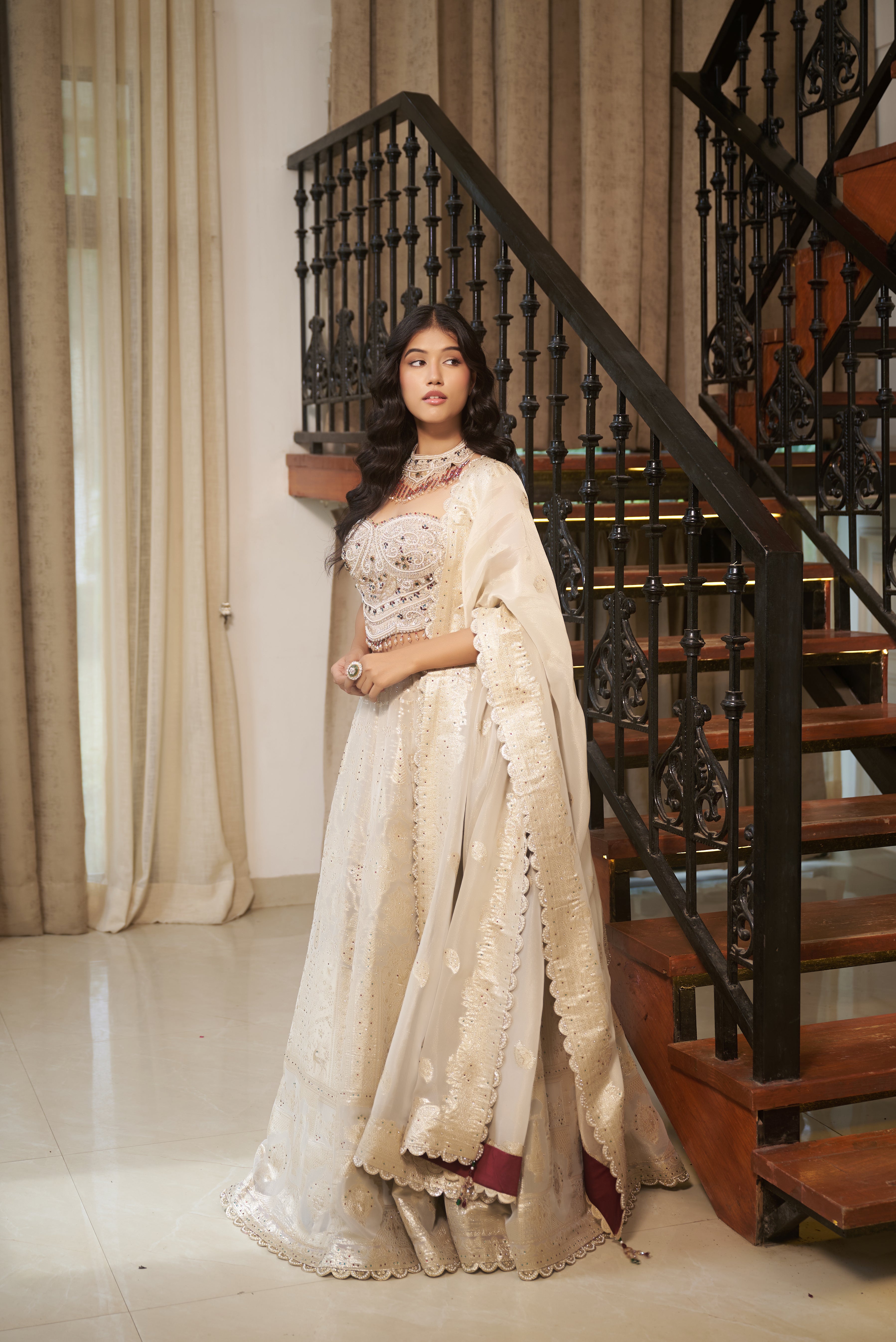 A woman wearing a traditional Indian outfit with a cream-colored embroidered kurta and a matching dupatta, standing in front of an indoor staircase Pista Floral Jaal Stone & Pearl Lehenga Set by MeenaGurnam